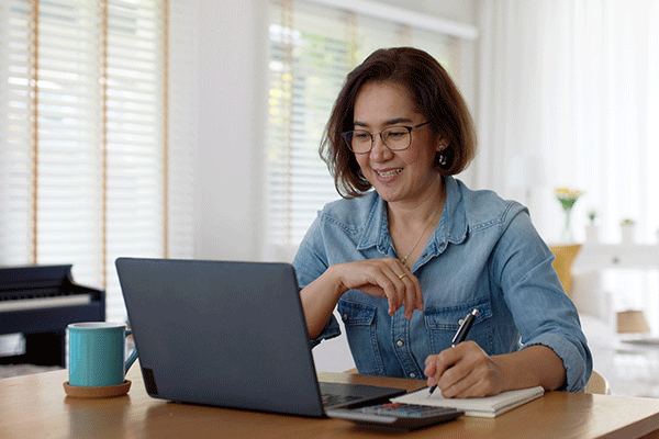 Middle-aged female of Asian descent with short brown hair, glasses, and wearing a denim shirt while sitting at a table with a laptop and writing on a notepad with her right hand. A calculator is visible on the table.  